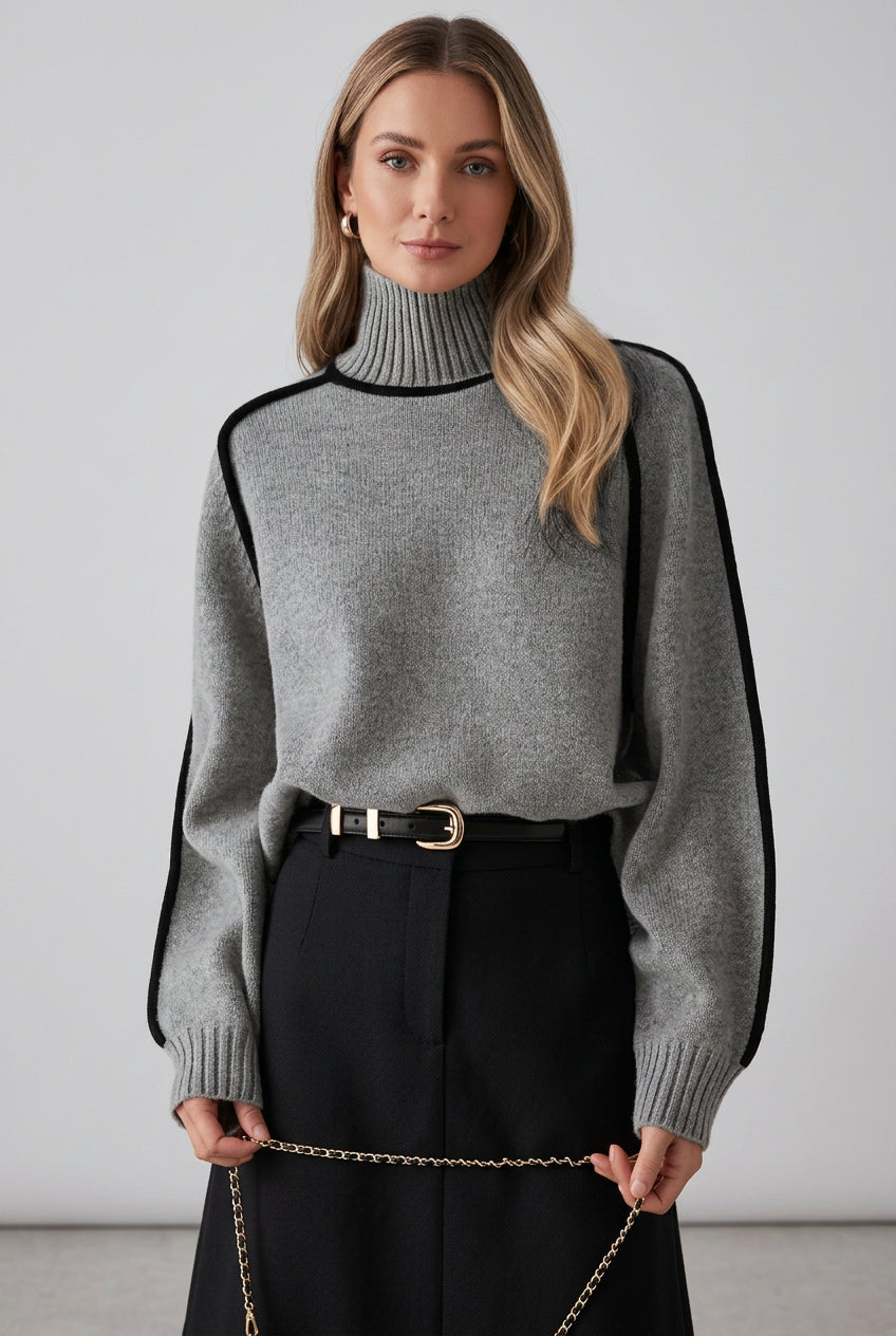 Person wearing a gray sweater with black accents and dark pants against a brick wall.