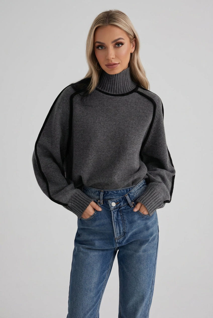 Person wearing a dark gray sweater with black accents and blue jeans against a brick wall.