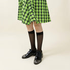 A person wearing a bright green and black plaid skirt, black knee-high socks, and shiny black-and-white patterned lace-up shoes, standing on white pebbles outdoors.