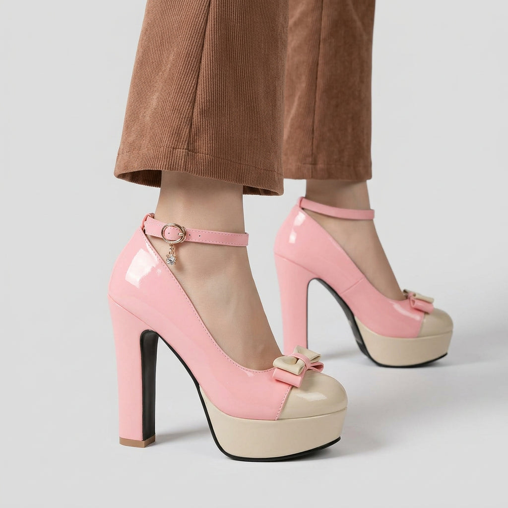 A pair of women's high-heeled platform sandals in light pink, shown being worn with brown pants on a wooden floor. The shoes feature a chunky block heel, a cream-colored platform sole, an ankle strap with a gold buckle, and a small bow detail on the toe.