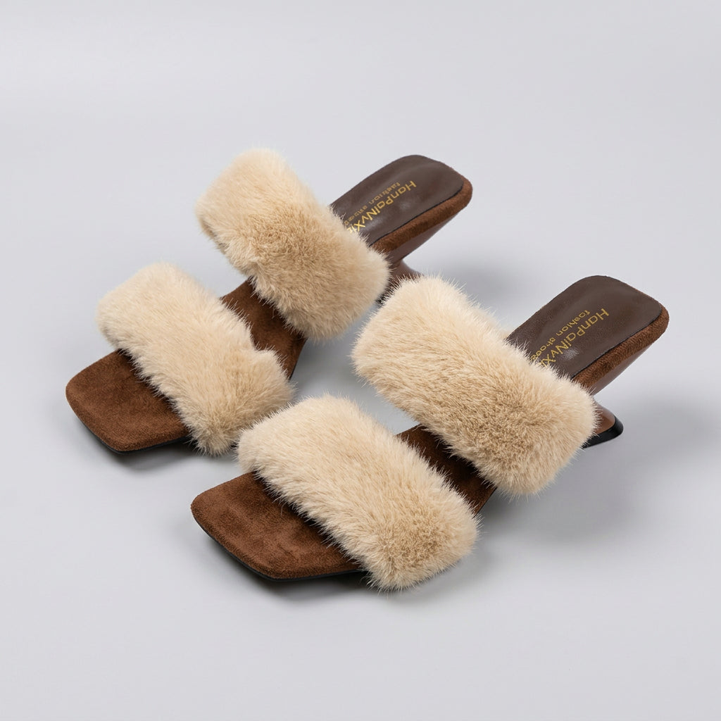 A pair of women's heeled slide sandals with thick beige faux-fur straps and brown suede-like square toes and heels, shown on a light gray floor with a mirror and bag in the background.