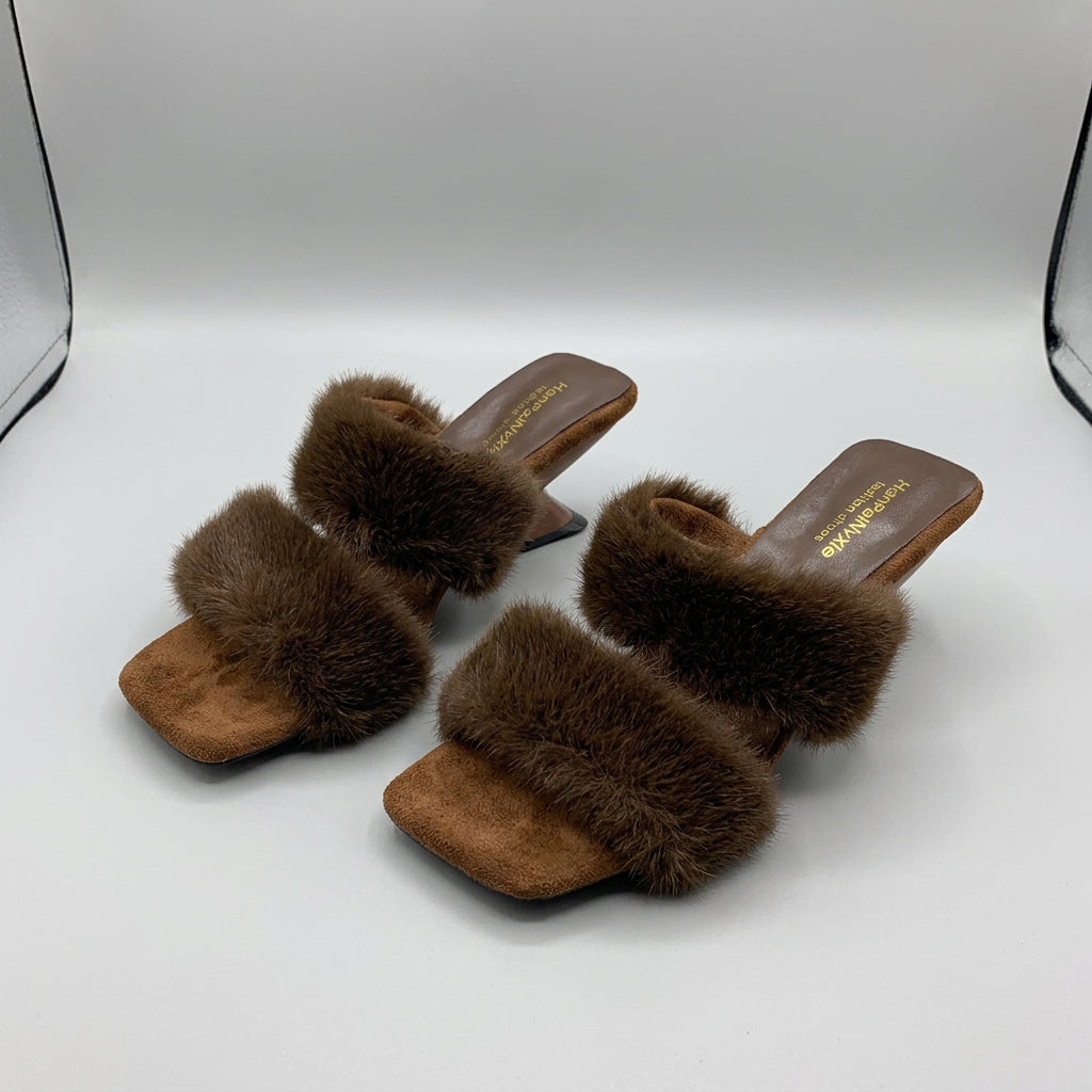 A pair of women's square-toe mule sandals with wide brown faux-fur straps and low block heels, shown on a light surface with a mirror and bag in the background.