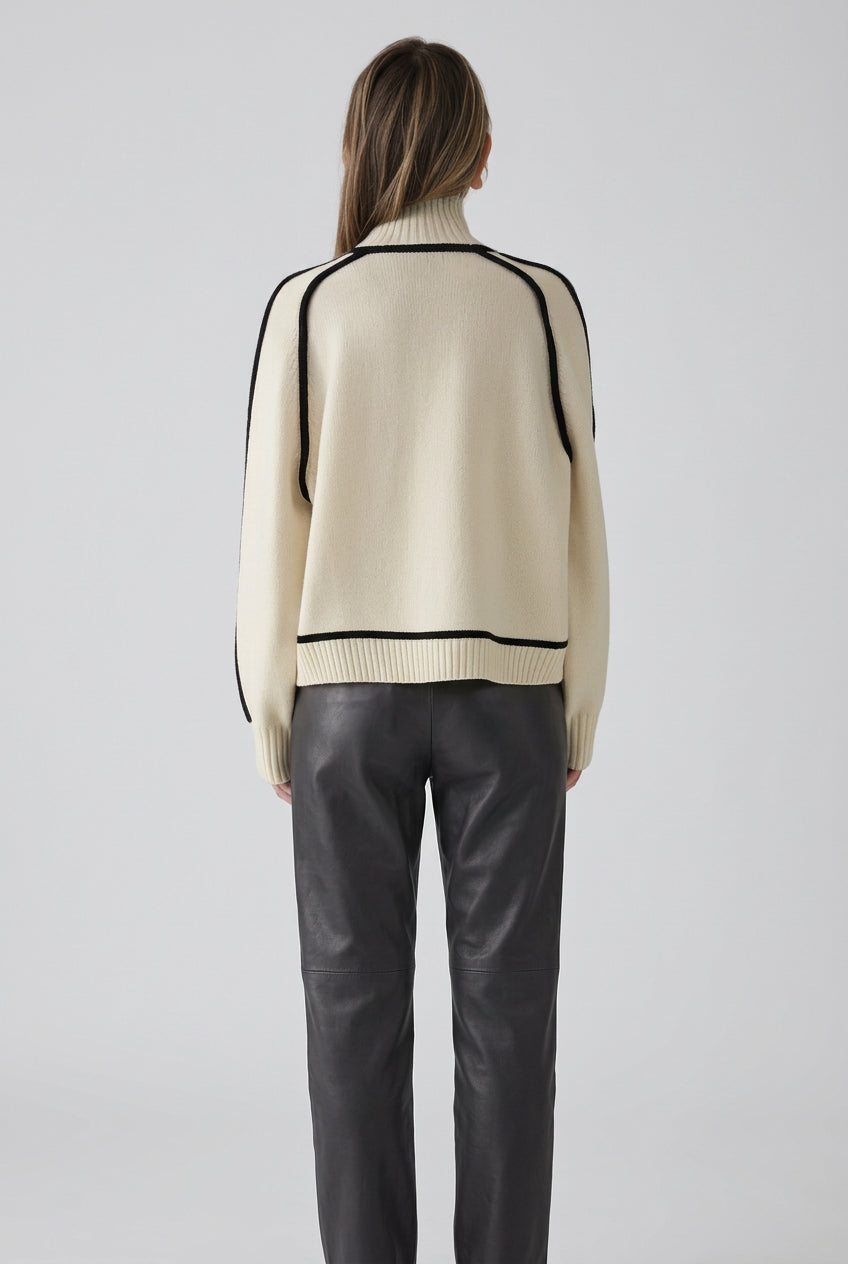Person wearing a cream sweater with black trim against a brick wall.