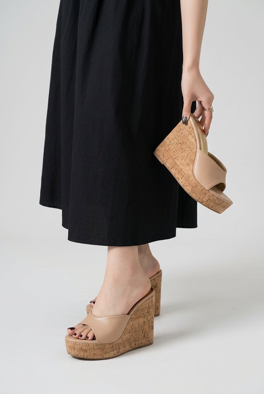 A person wearing a black dress sits on a bench, showing a pair of beige open-toe wedge sandals with cork-look platform soles. One shoe is worn on the foot, and the other is held in the hand to show the side profile.